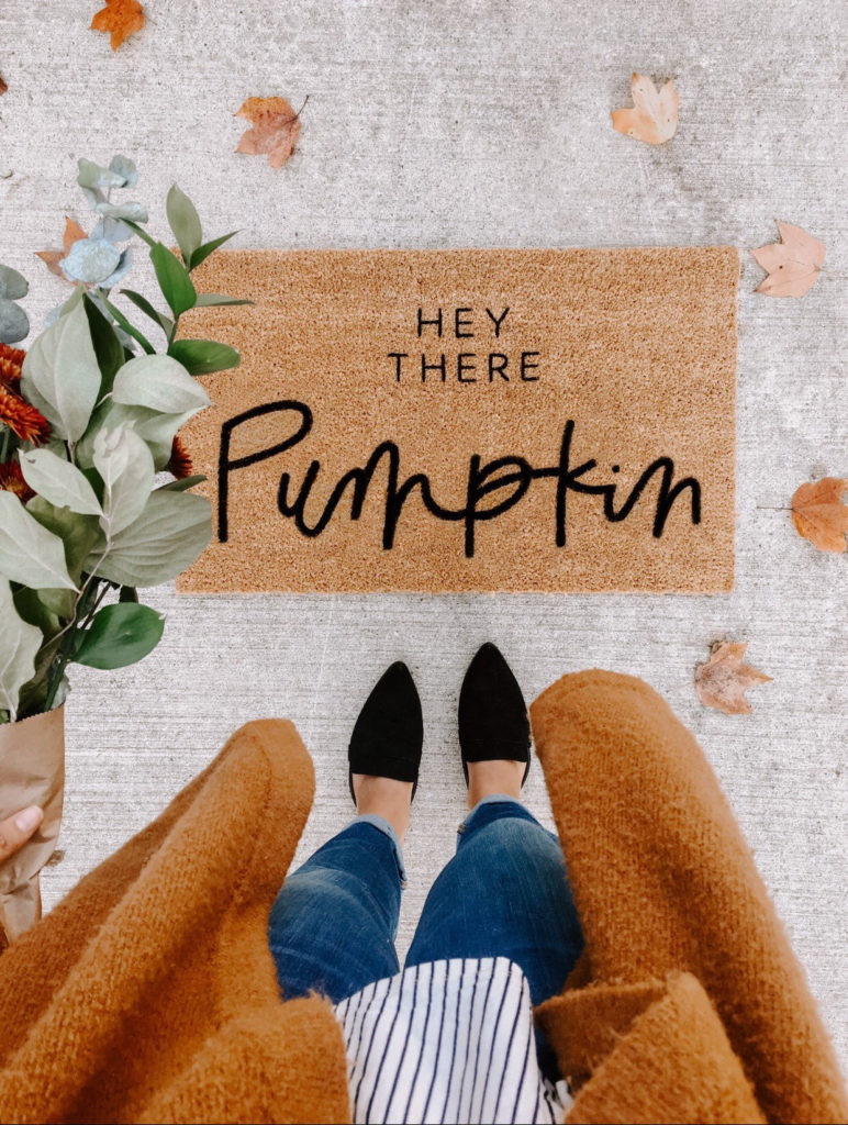 yellow-orange welcome mat that says hey there pumpkin, surrounded by autumn leaves and a pair of women's black shoes